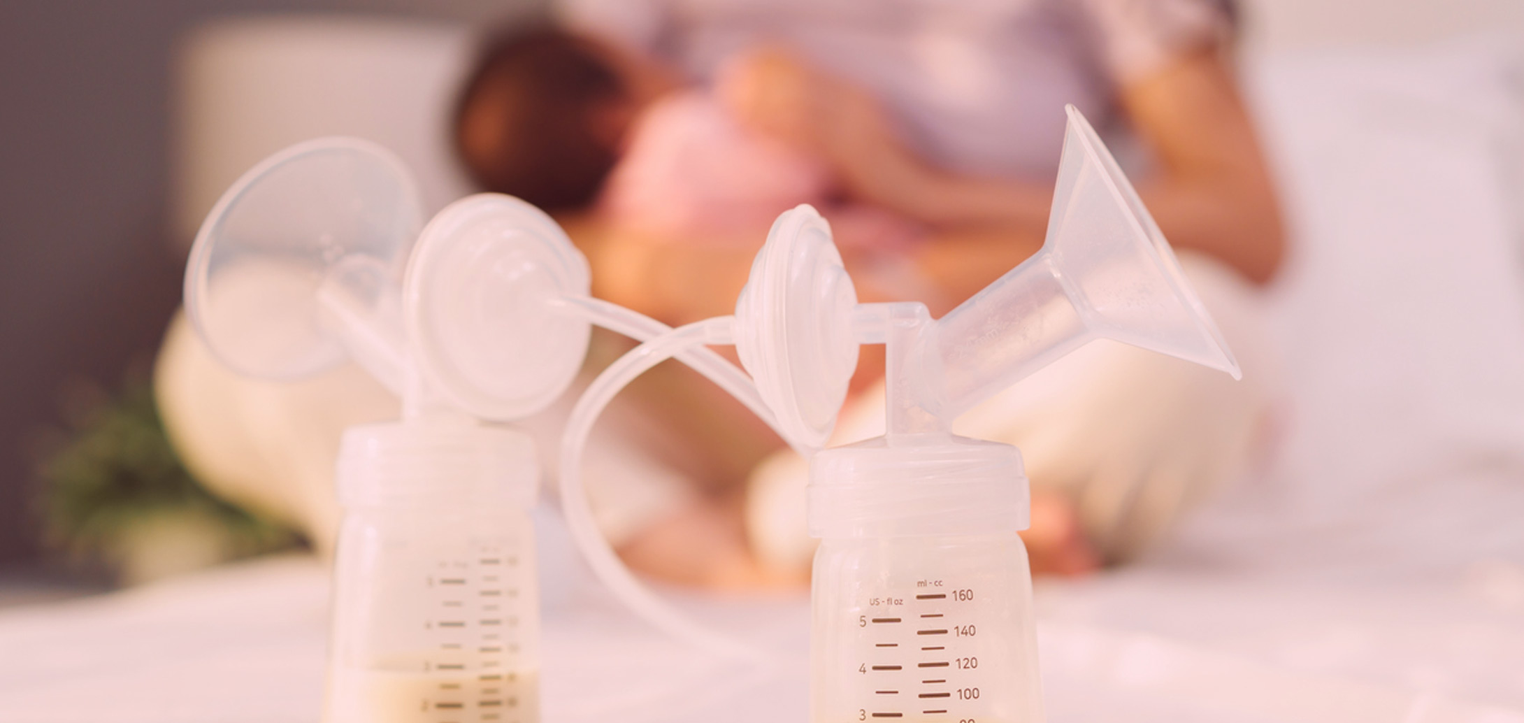 Two breast pumps in foreground, nursing mother sitting out of focus in background